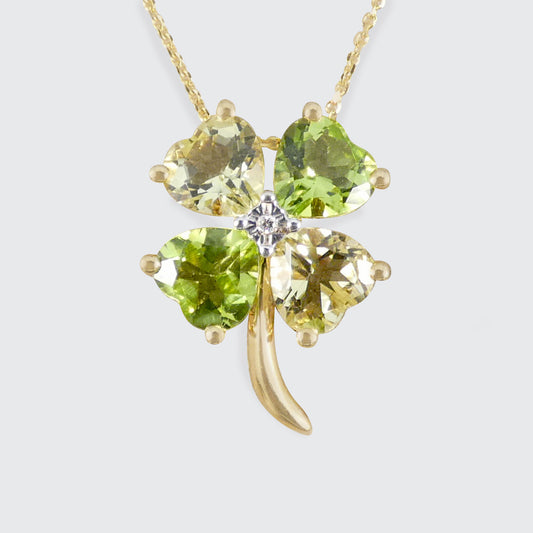 A yellow gold four-leaf clover pendant necklace with four faceted green gemstones and a small clear center stone, shown on a light gray background.