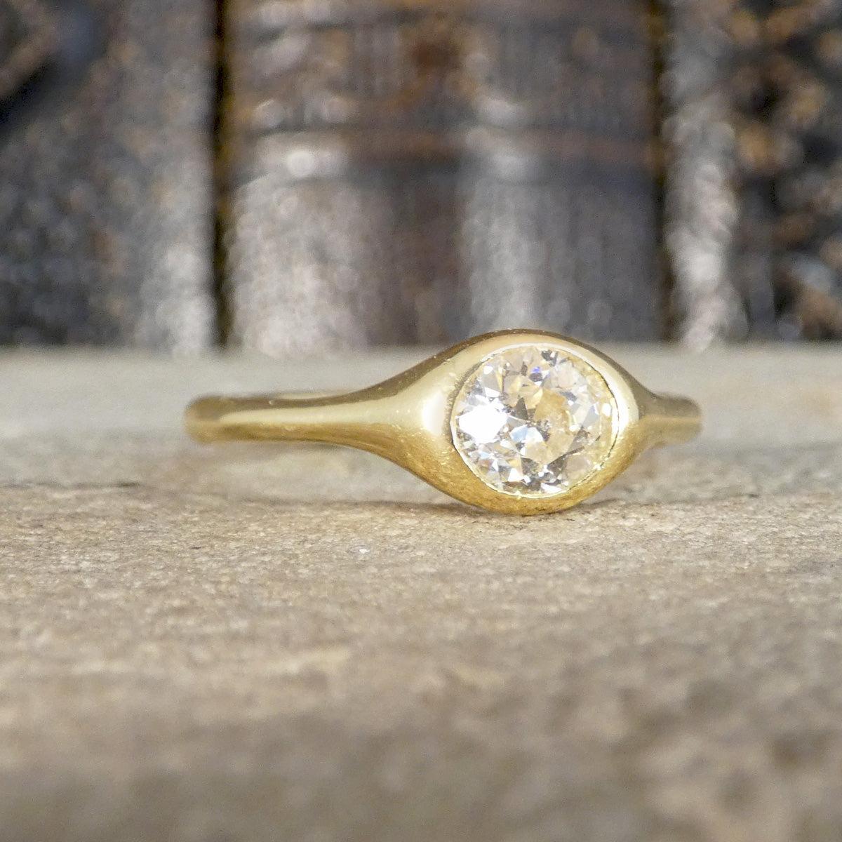 A gold ring with a round clear gemstone set in a bezel-like oval, resting on a textured stone surface with a blurred background.