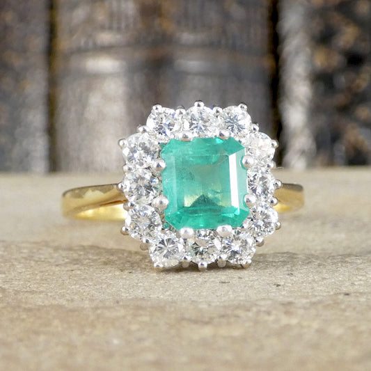 A yellow gold ring featuring a square-cut emerald-cut green gemstone surrounded by a double halo of clear round-cut stones, displayed on a textured stone surface with a blurred background.