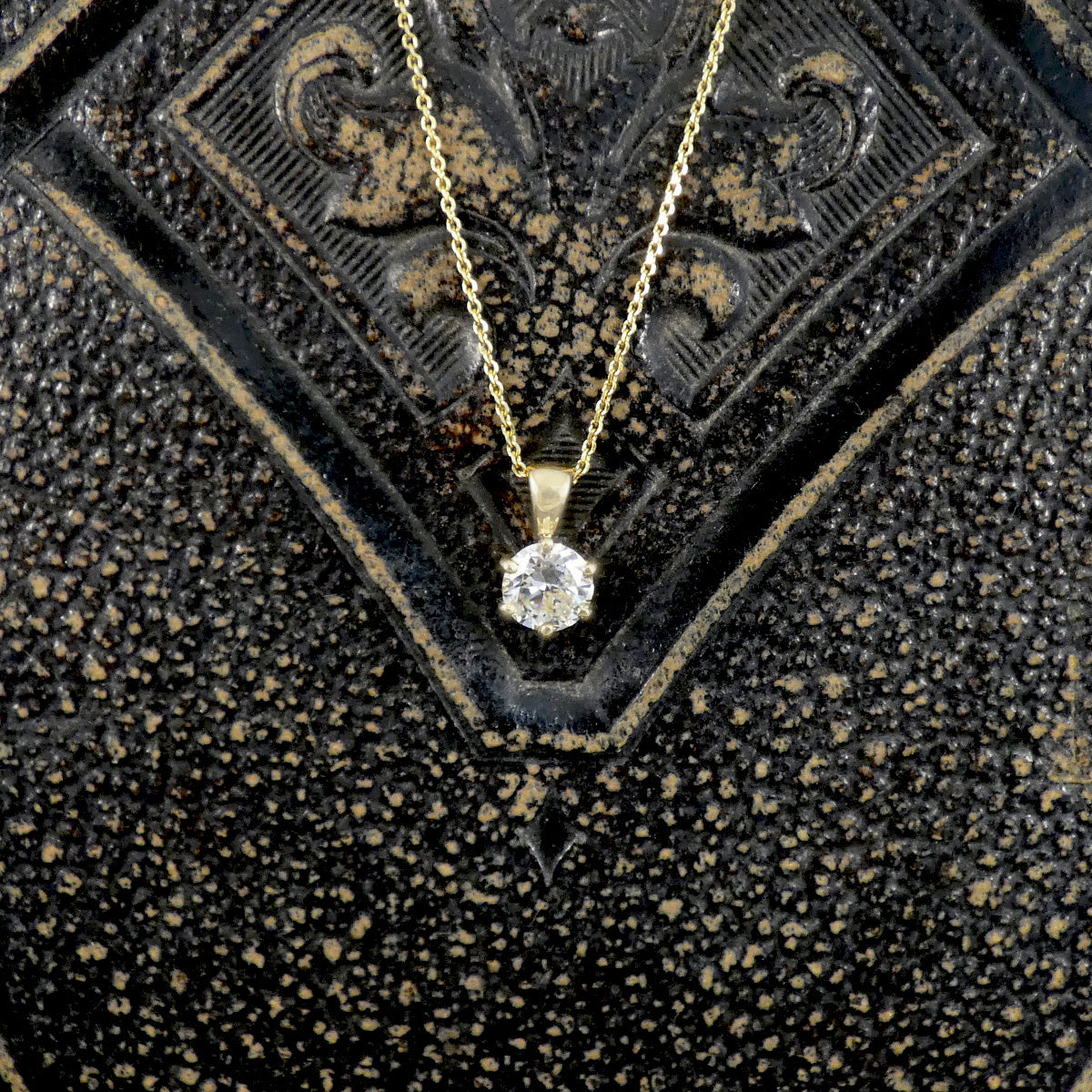 A gold necklace with a round-cut clear diamond gemstone pendant, displayed on a textured black and gold decorative surface.