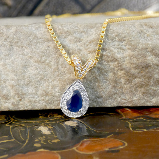 A gold chain necklace with a teardrop-shaped pendant featuring a deep blue center stone surrounded by small clear stones, displayed on a textured stone block.
