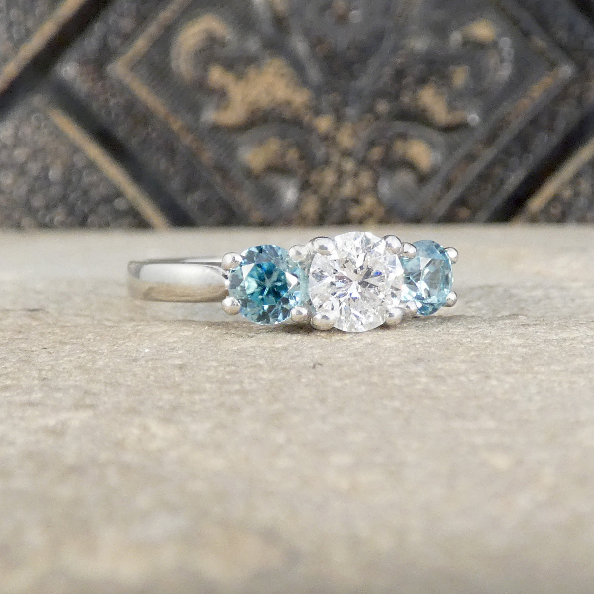 A platinum three-stone ring featuring a round brilliant-cut center diamond flanked by two oval light blue zircons, set in a platinum band.