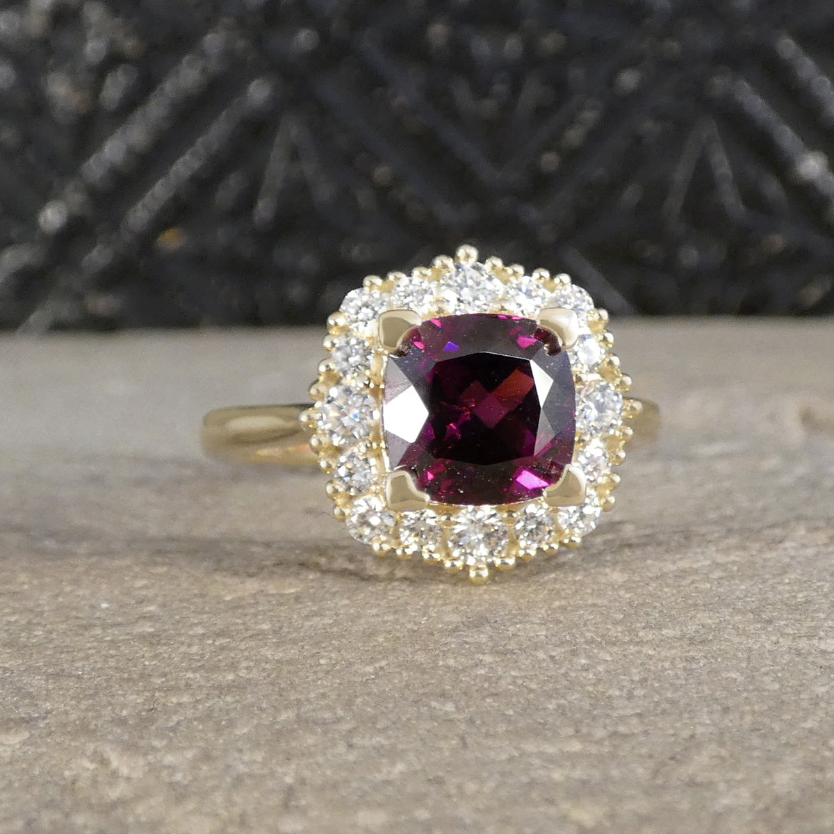 A yellow gold ring featuring a cushion-cut deep magenta-purple center gemstone surrounded by a double halo of small clear stones, resting on a textured gray stone surface.