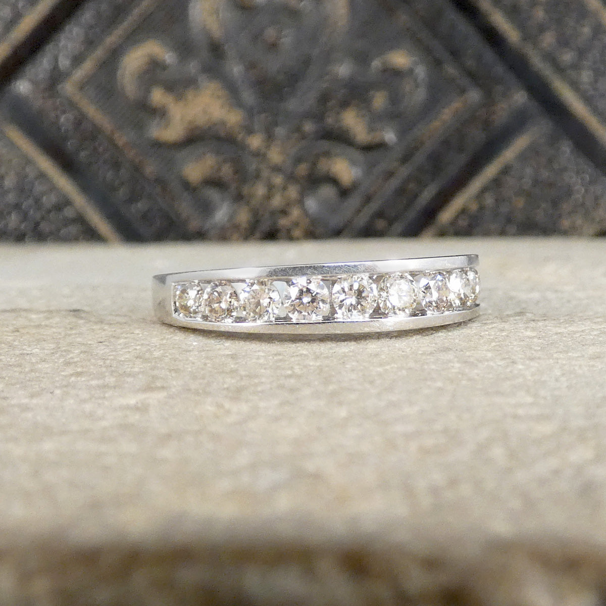 A silver band ring featuring a row of clear round-cut gemstones, displayed on a textured beige surface with a dark, ornate background.