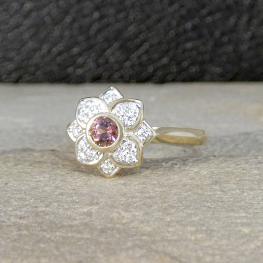 Gold ring with a central pink gemstone and diamond-studded petals on a textured surface.