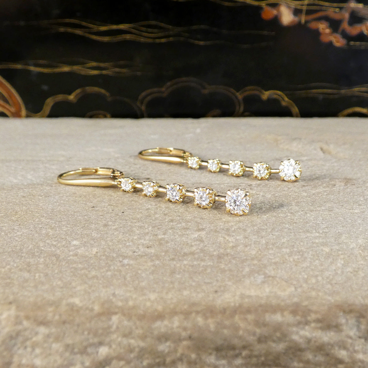 A pair of gold leverback earrings with a row of clear round-cut gemstones, displayed on a textured beige stone surface with a dark, patterned background.