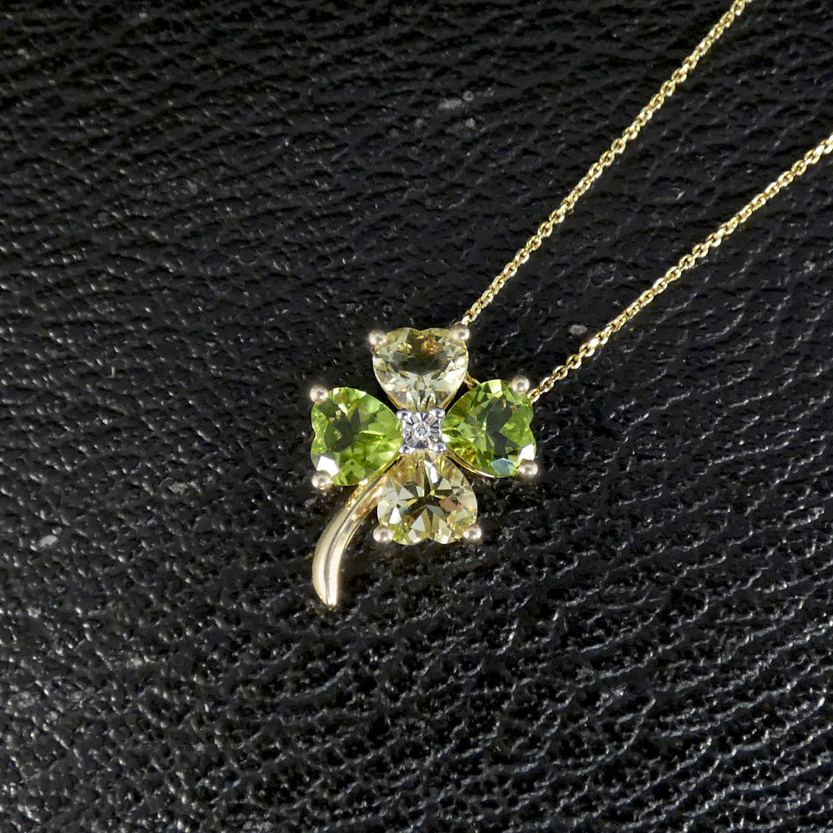 A gold necklace with a four-leaf clover pendant made of green gemstones and a small clear center stone, displayed on a textured black background.