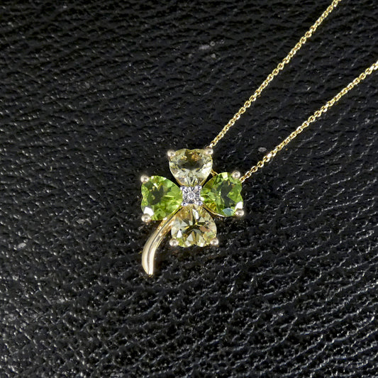 A gold necklace with a four-leaf clover pendant made of green gemstones and a small clear center stone, displayed on a textured black background.