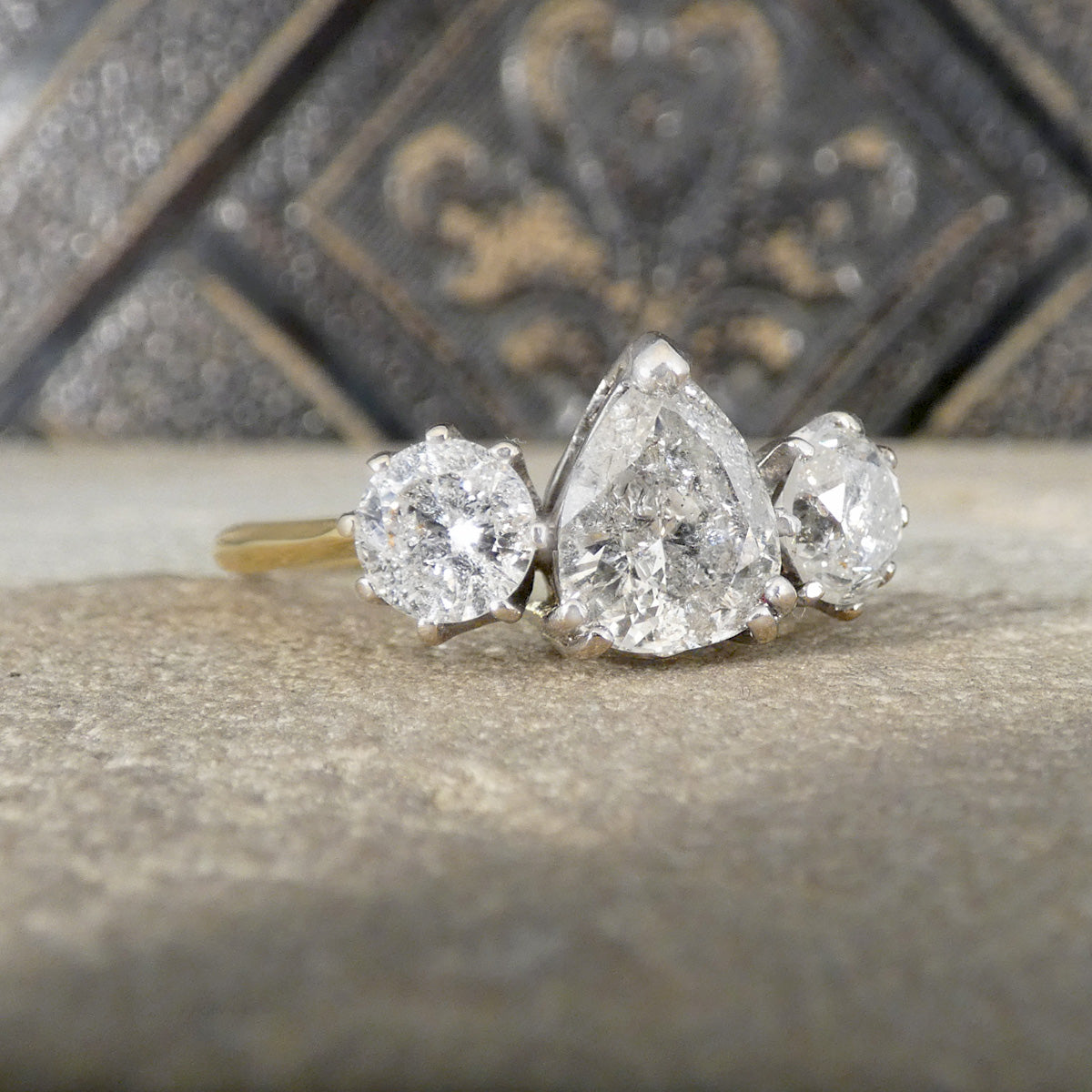 Three-stone diamond ring with pear-shaped center stone on a textured surface.