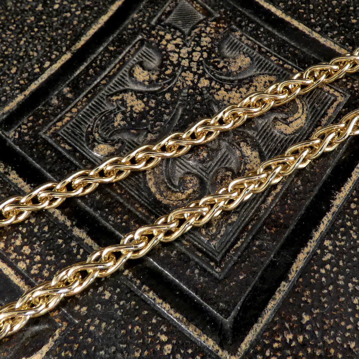 A close-up of a polished gold chain draped diagonally across a dark, textured embossed surface.
