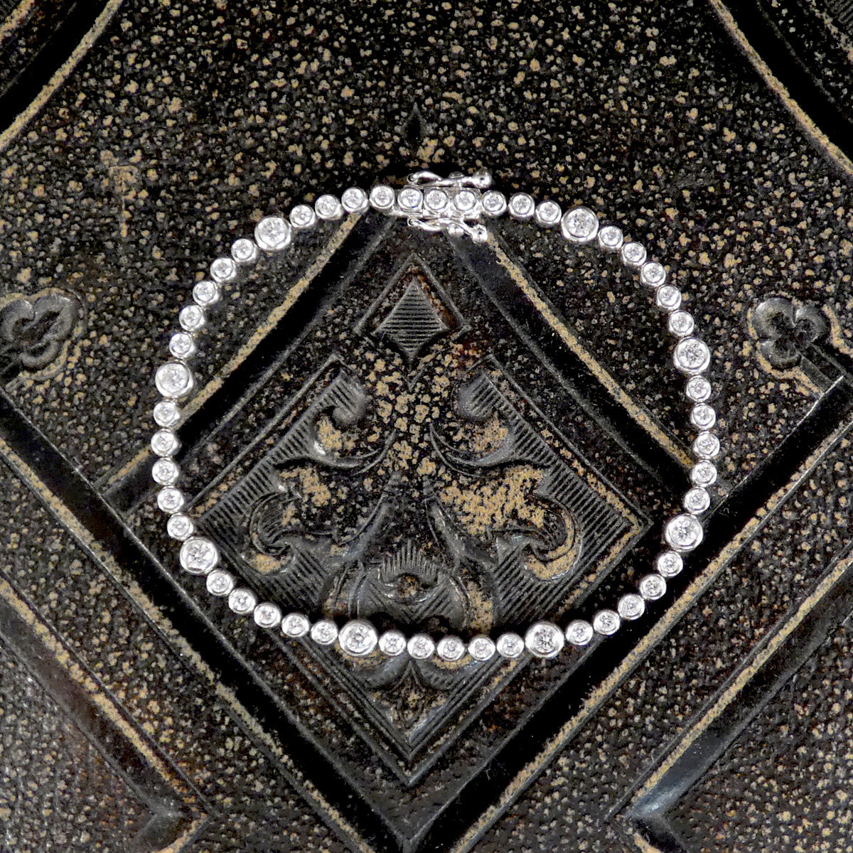 A diamond tennis bracelet with bezel set diamonds of different sizes in white metal on a dark background.