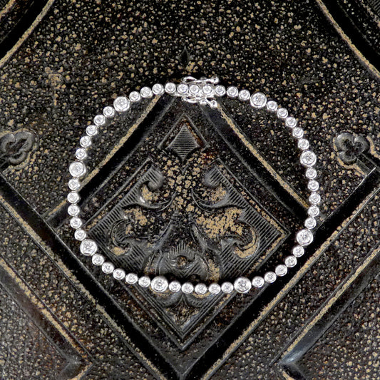 A diamond tennis bracelet with bezel set diamonds of different sizes in white metal on a dark background.
