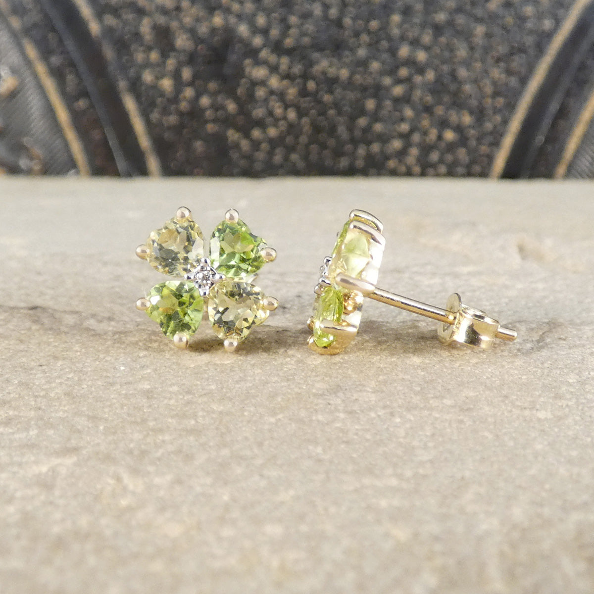 A pair of yellow gold stud earrings with four green gemstones arranged in a flower shape, resting on a textured beige stone surface with a dark patterned background.