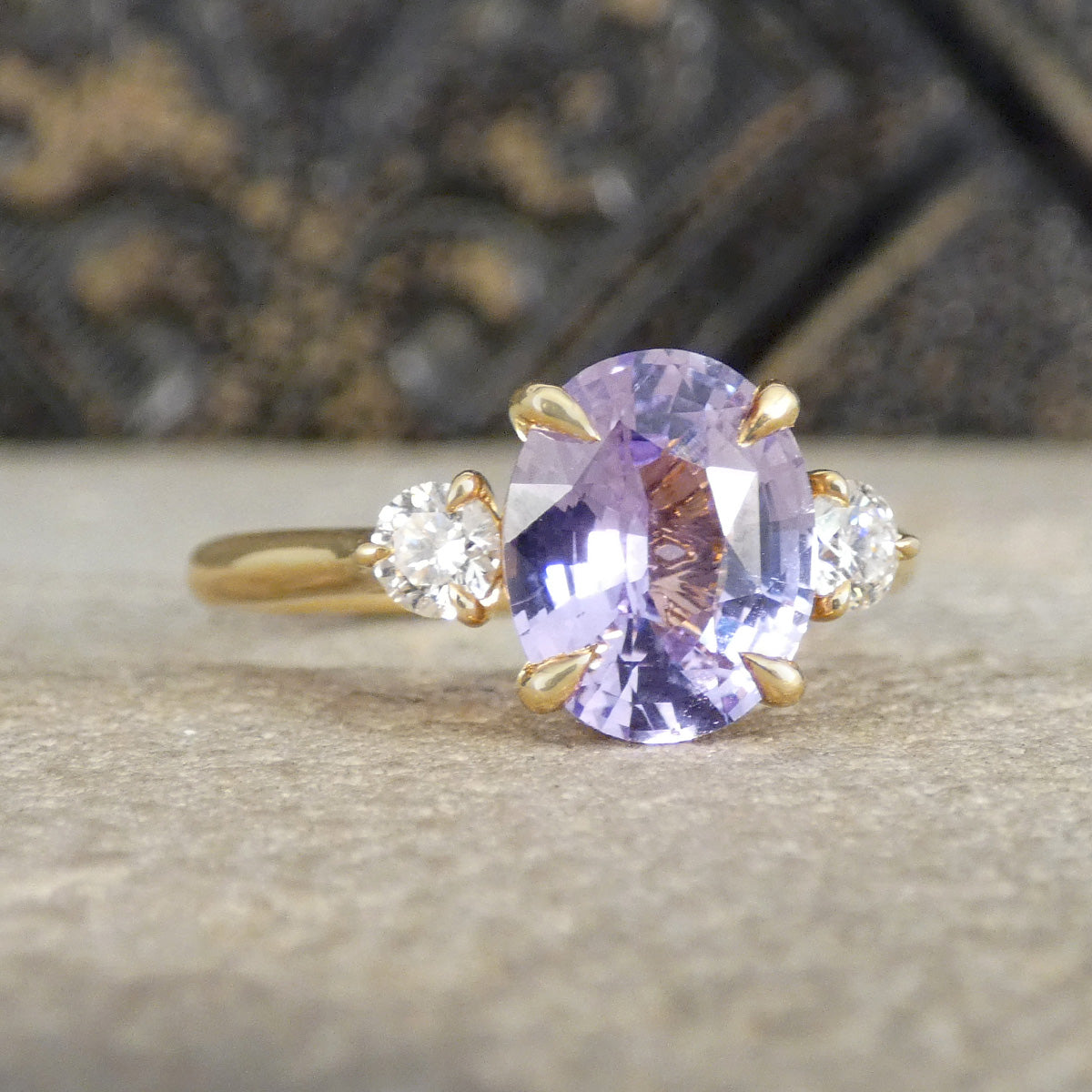 A yellow gold ring featuring a large oval-cut light purple sapphire center stone and two small round clear accent stones on either side, set against a textured gray stone background.