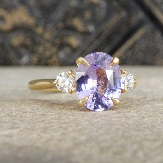 A yellow gold ring featuring a large oval-cut light purple sapphire center stone and two small round clear accent stones on either side, set against a textured gray stone background.