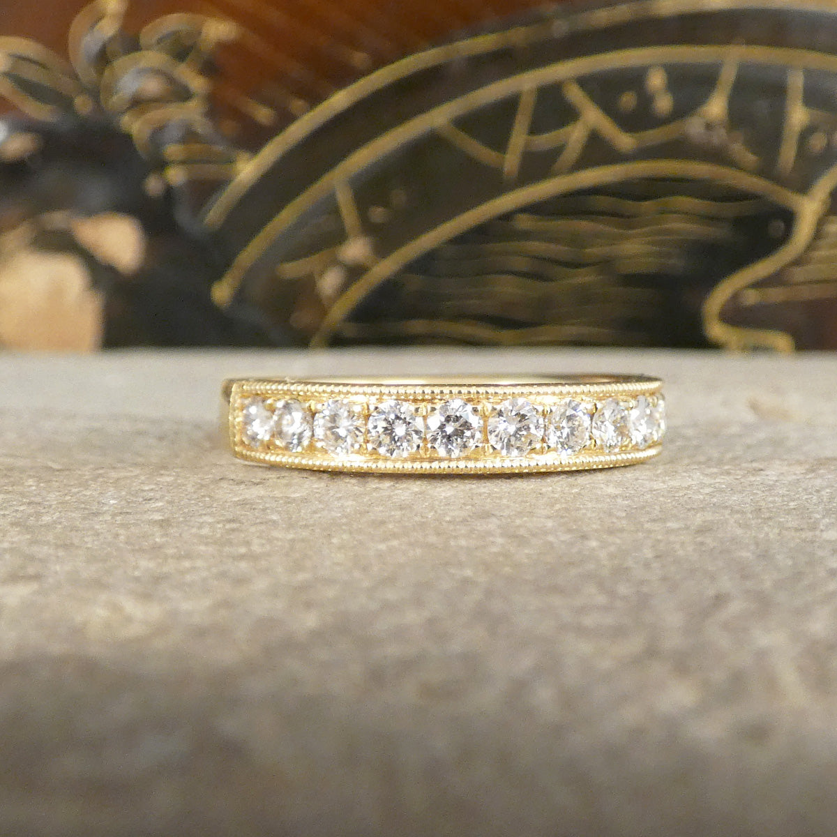 A yellow gold eternity ring featuring a row of round-cut clear diamond gemstones, displayed on a textured gray stone surface with a blurred decorative background.
