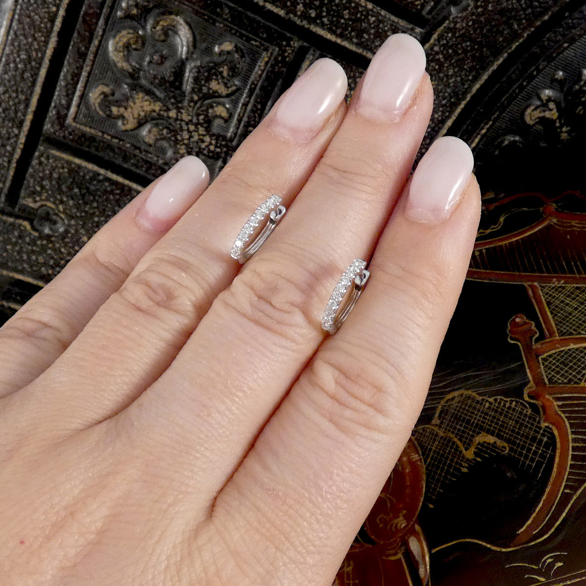 A close-up of a hand with pale pink manicured nails wearing two small silver huggie hoop rings set with clear pavé stones, posed against a dark ornate background.