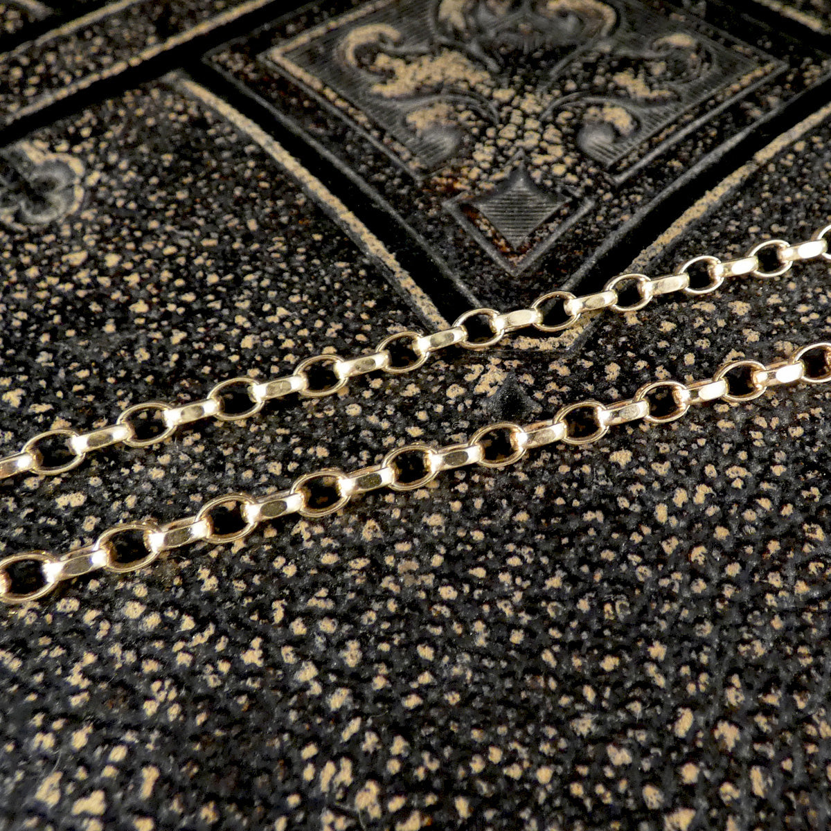 A close-up of a gold chain with elongated oval links laid diagonally across a textured black and gold patterned surface.