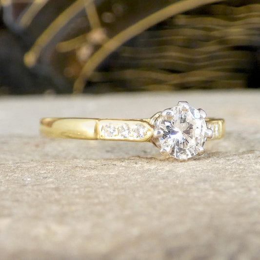 A yellow gold solitaire engagement ring with a round-cut diamond center stone and small pavé diamonds on the band, resting on a textured stone surface.