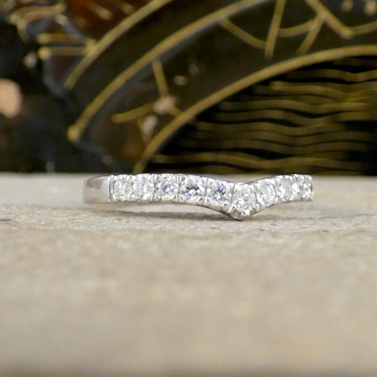 A silver band ring featuring a wavy, chevron-like arrangement of clear pavé-set gemstones, resting on a light beige textured surface with a dark, patterned background.
