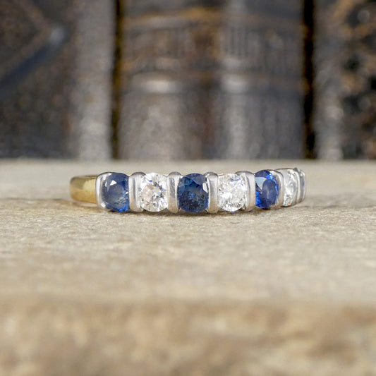 A gold band ring featuring a row of alternating round blue sapphire and clear round diamonds, resting on a textured stone surface with a blurred background of antique books.