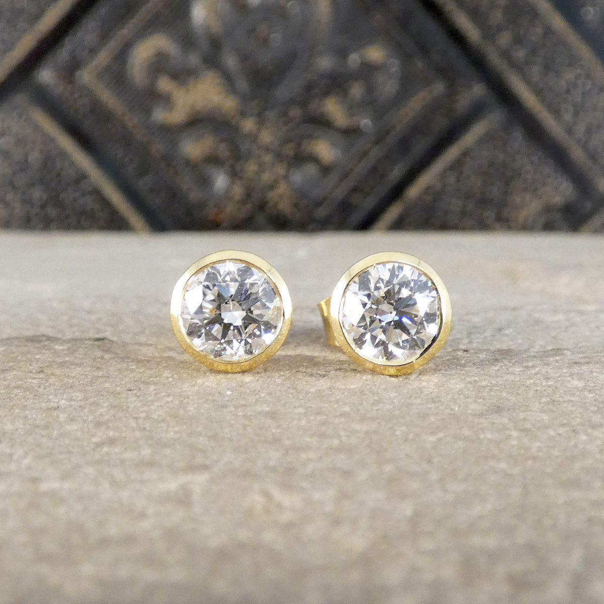 A pair of round gold stud earrings with clear faceted gemstones, displayed on a textured stone surface with a dark decorative background.