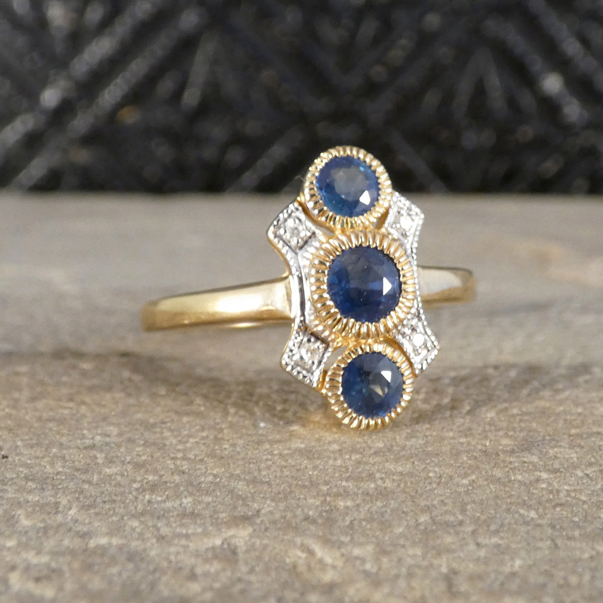 A gold ring with three oval blue sapphires and small clear accent stones, displayed on a textured stone surface with a dark patterned background.