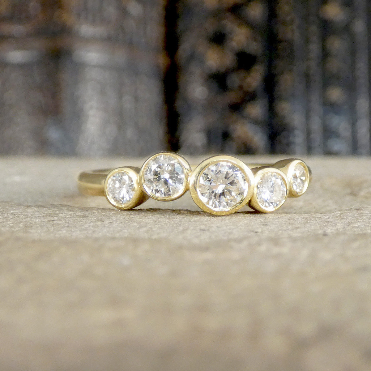 A yellow gold ring featuring four round bezel-set clear gemstones of varying sizes, displayed on a textured stone surface with a blurred dark background.