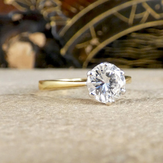 A classic solitaire engagement ring featuring a round brilliant-cut diamond set in a yellow gold band, resting on a textured stone surface with a blurred decorative background.