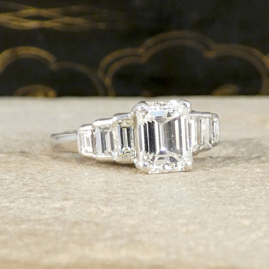 An emerald-cut diamond ring with baguette side stones in a silver-toned setting, resting on a textured beige stone surface with a dark, patterned background.