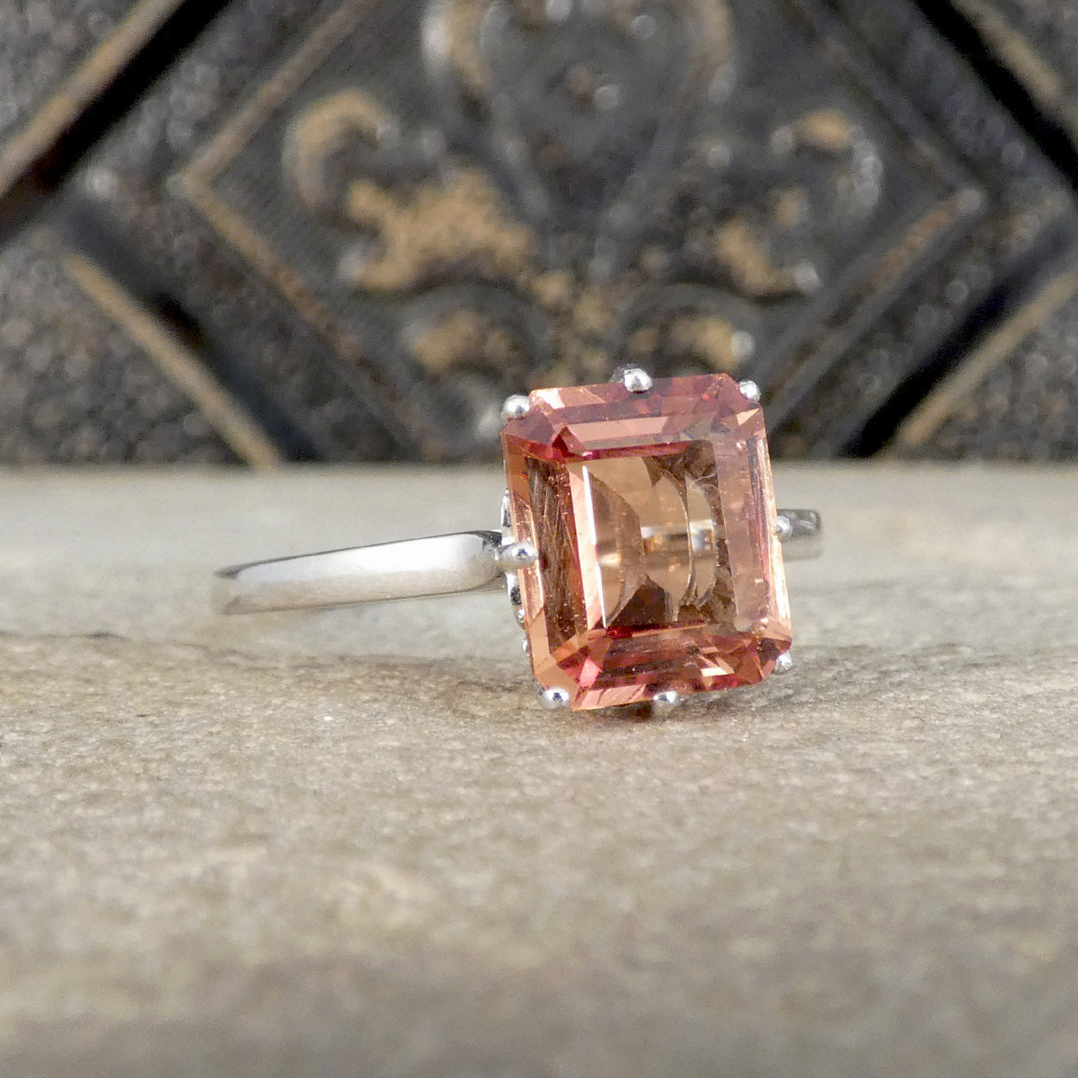 A silver ring featuring a rectangular peach-colored gemstone, resting on a textured stone surface with a dark, ornate background.
