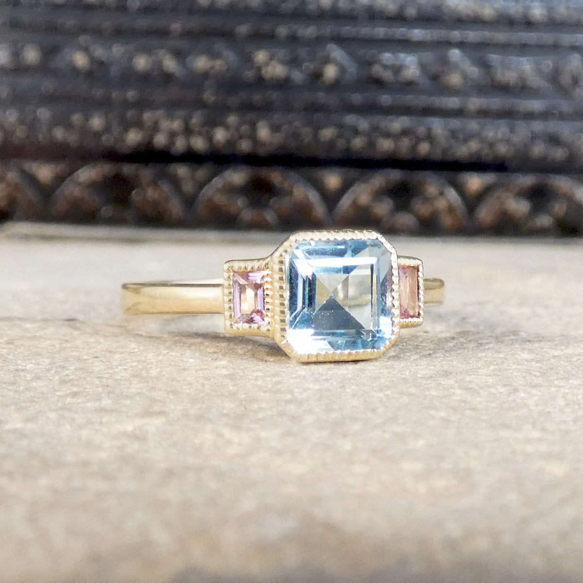 A gold ring featuring a large emerald-cut light blue center stone and two small pink  side stones, displayed on a textured beige surface with a dark carved wooden background.