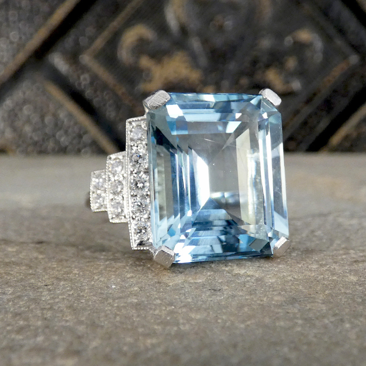 An emerald-cut light blue aquamarine ring set in silver with diamond accents on a textured stone surface.