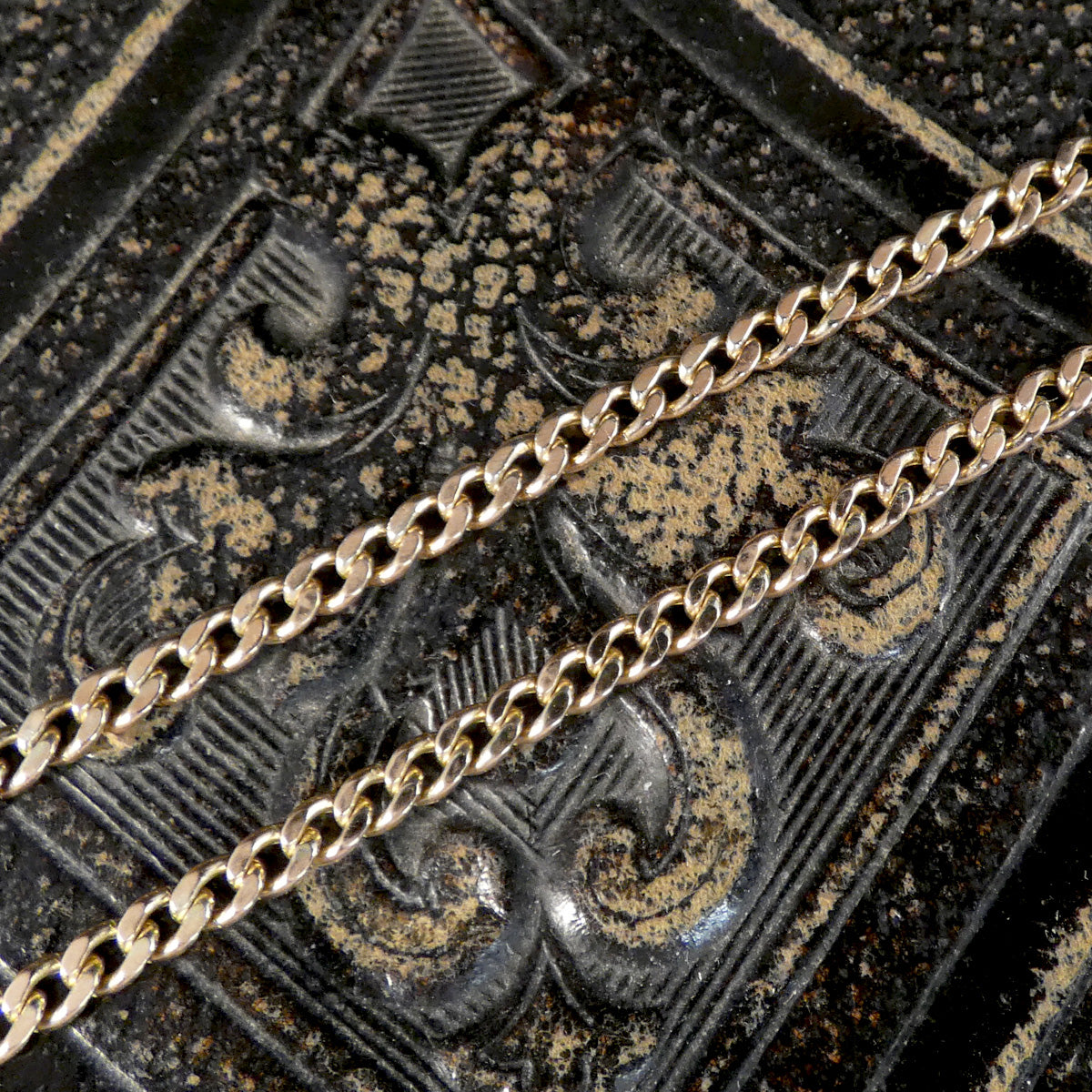 Two gold-toned metal chains laid diagonally across a dark, ornate textured surface with raised decorative patterns.