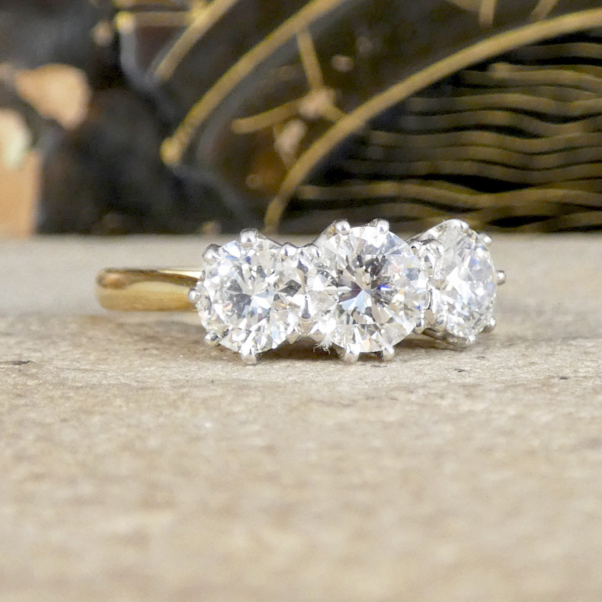 A three-stone diamond ring with a yellow gold band, featuring three round brilliant-cut diamonds set in a row, resting on a textured stone surface with a blurred background.