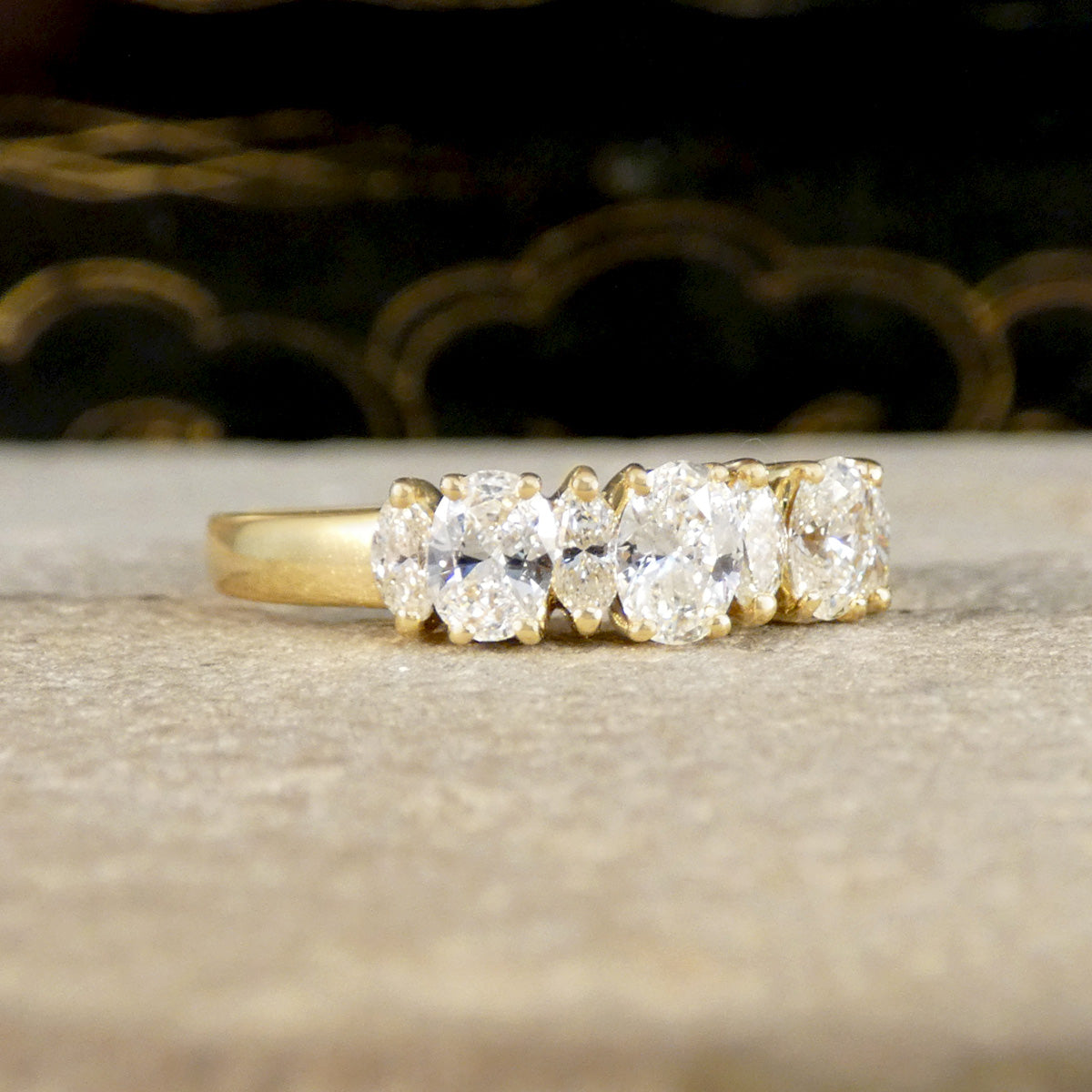 A yellow gold diamond ring with oval-cut diamonds, resting on a textured beige surface with a dark, blurred background.