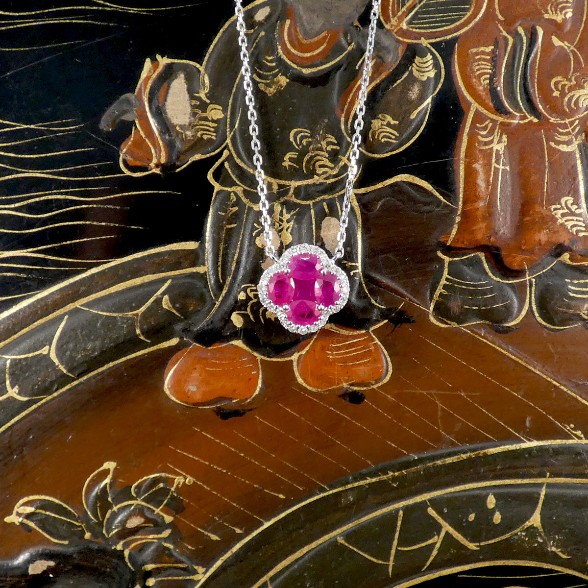 A bright pink ruby and diamond cluster necklace on an ornate background.