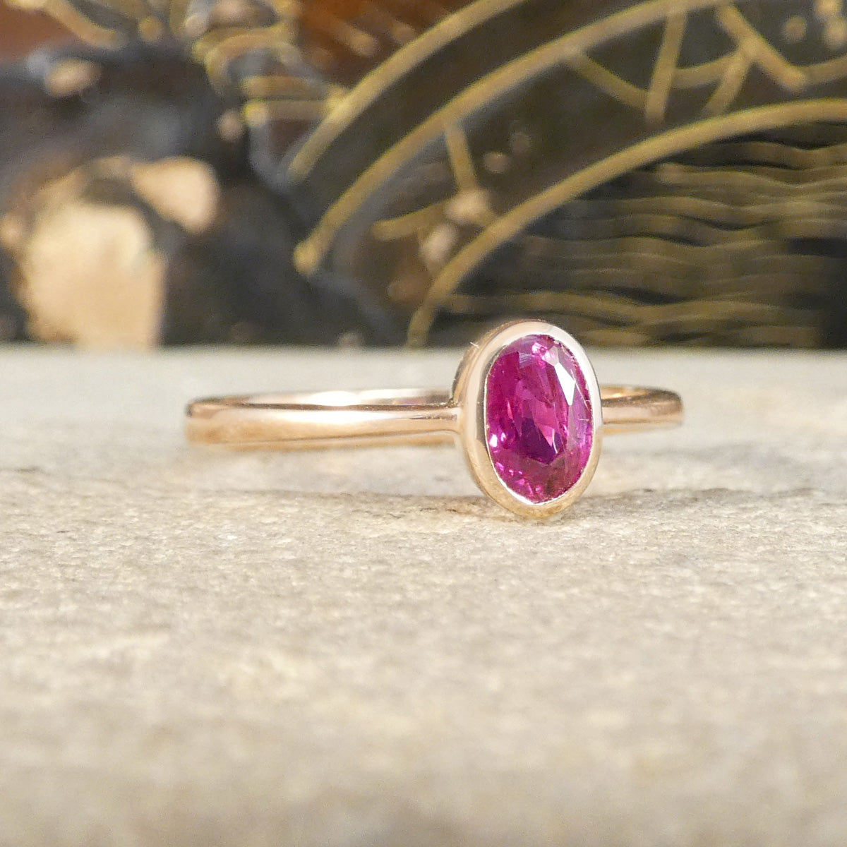 A gold ring with an oval-cut pinkish ruby, resting on a textured light beige surface with a blurred dark background.