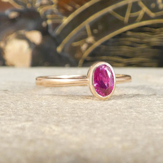 A gold ring with an oval-cut pinkish ruby, resting on a textured light beige surface with a blurred dark background.