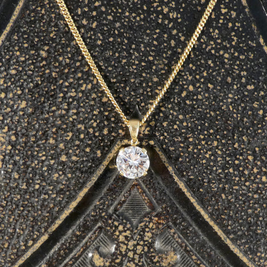A yellow gold necklace with a single round-cut clear diamond gemstone pendant, displayed on a textured black and gold geometric jewelry box.