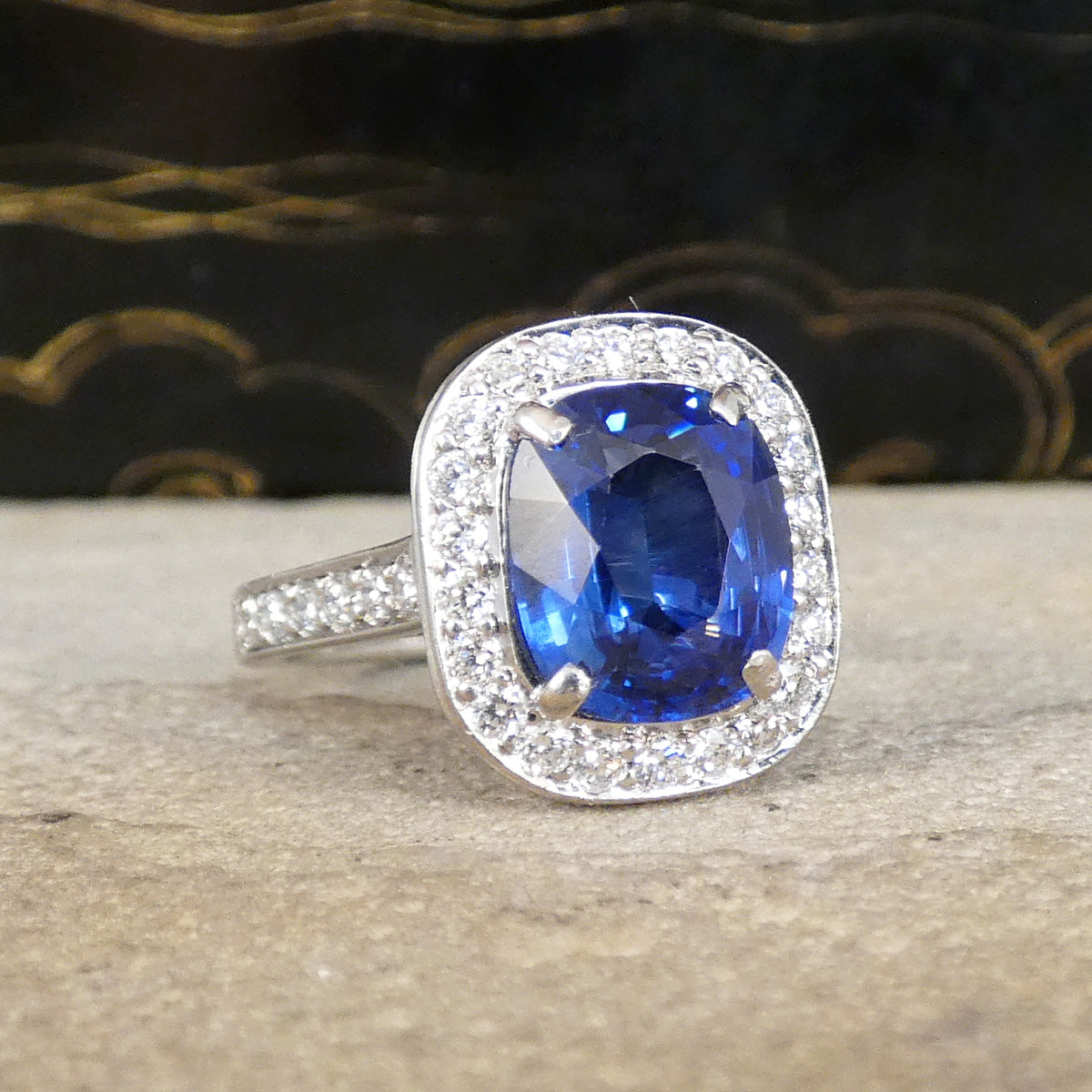 A cushion-cut blue sapphire ring with a diamond halo and a silver-colored band, resting on a textured stone surface with a dark patterned background.