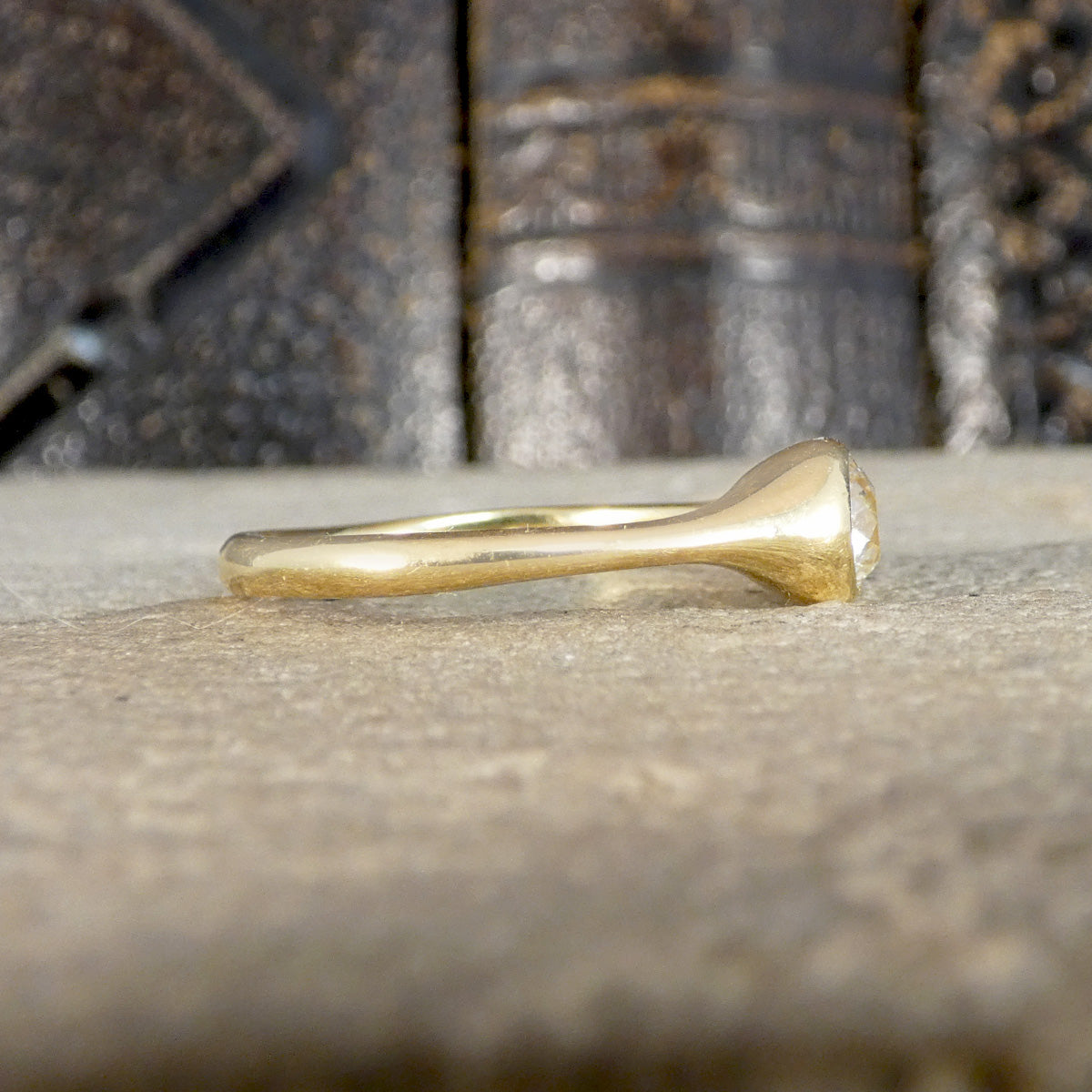 A gold ring with a unique mushroom-shaped setting, resting on a textured stone surface with a blurred background of dark books.
