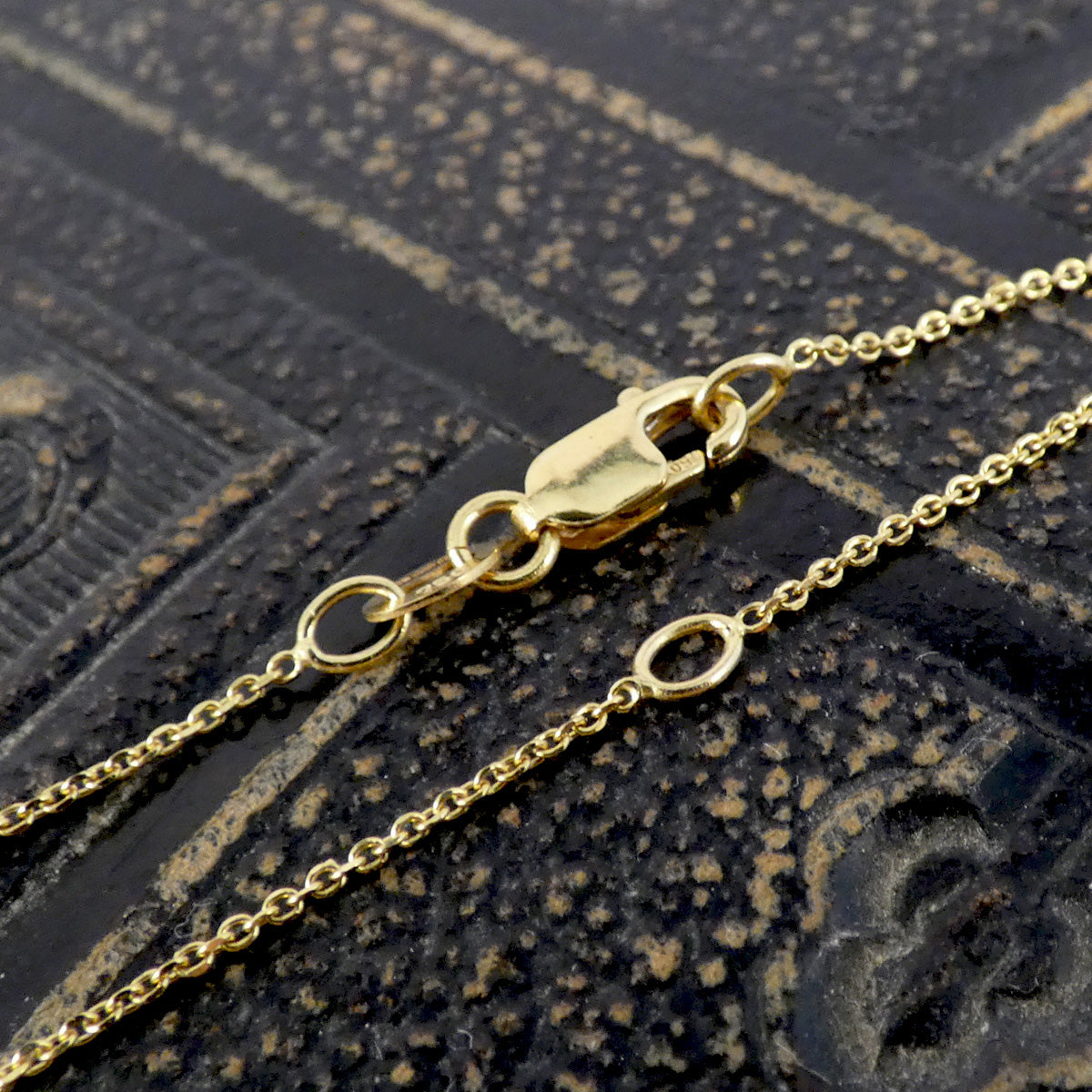 A close-up of a gold chain necklace with a lobster clasp and small circular links, laid on a dark textured surface.