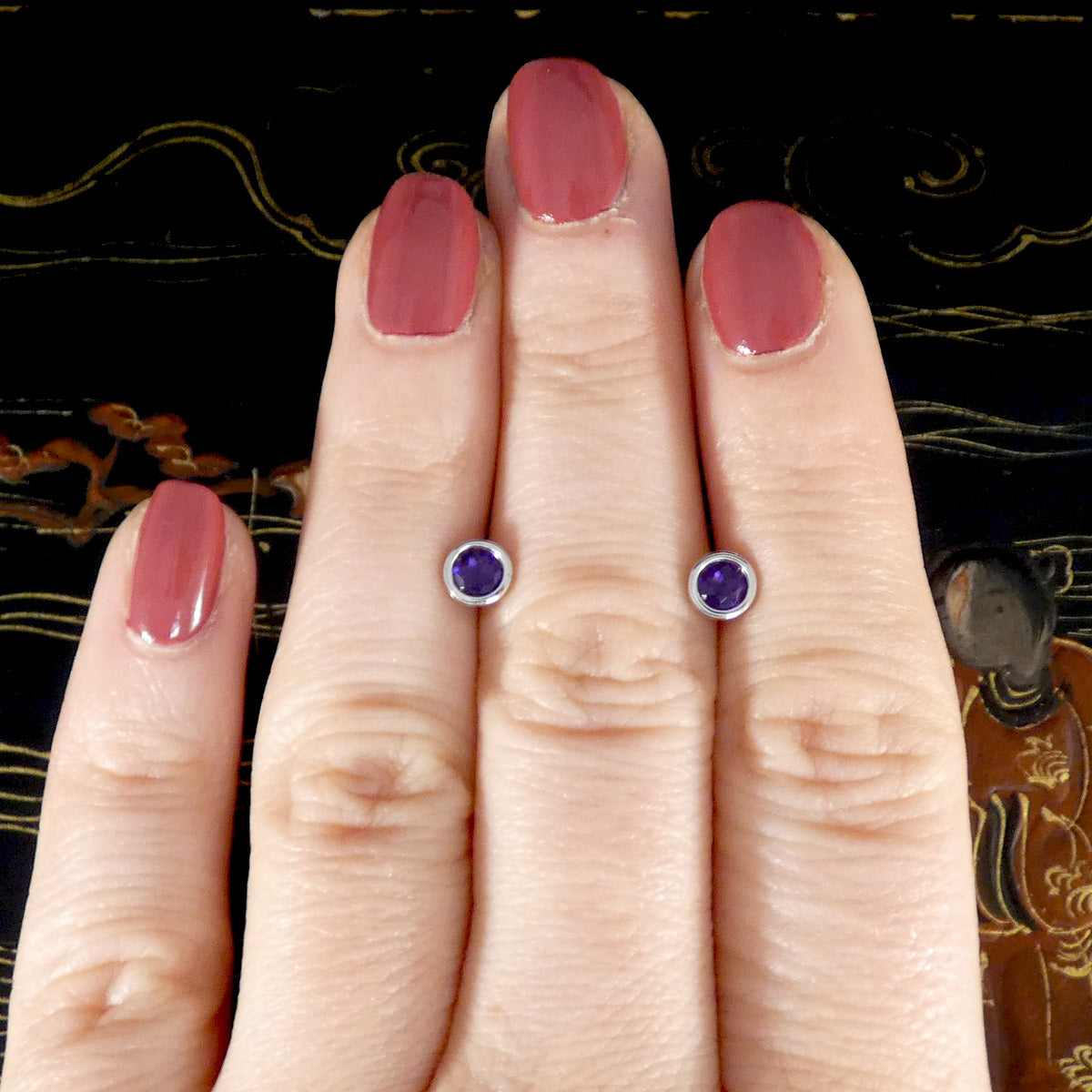 Hand wearing purple gemstone stud earrings with a decorative background
