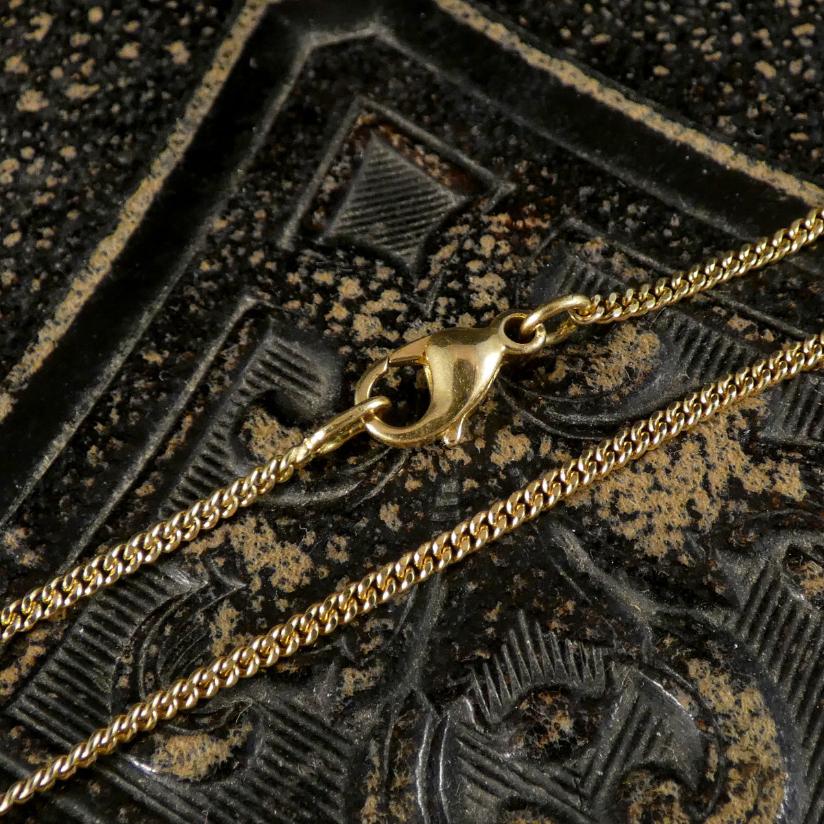 A close-up of a gold-toned chain necklace with a lobster clasp, laid diagonally across a dark textured surface with gold speckling.