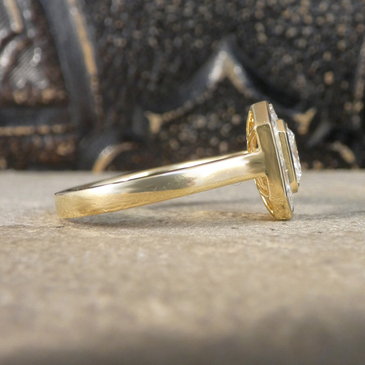 A close-up shot of a yellow gold ring with a geometric, bezel setting holding a clear gemstone, resting on a textured stone surface.