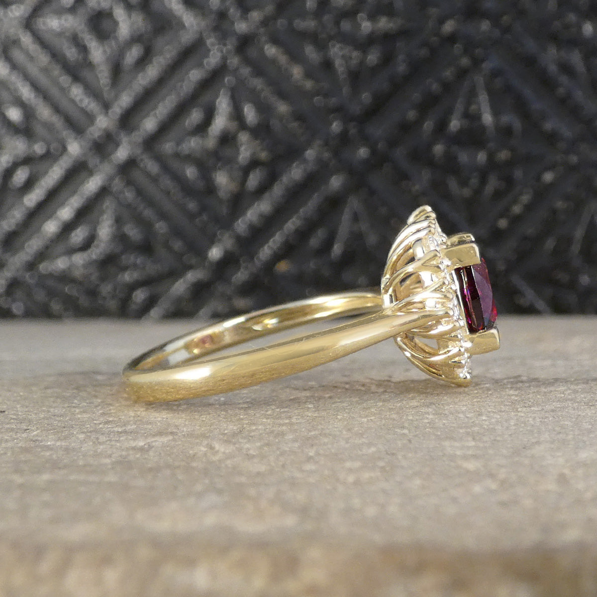A yellow gold ring with a textured, flame-like halo setting holding a rectangular dark red gemstone, resting on a light stone surface with a dark patterned background.