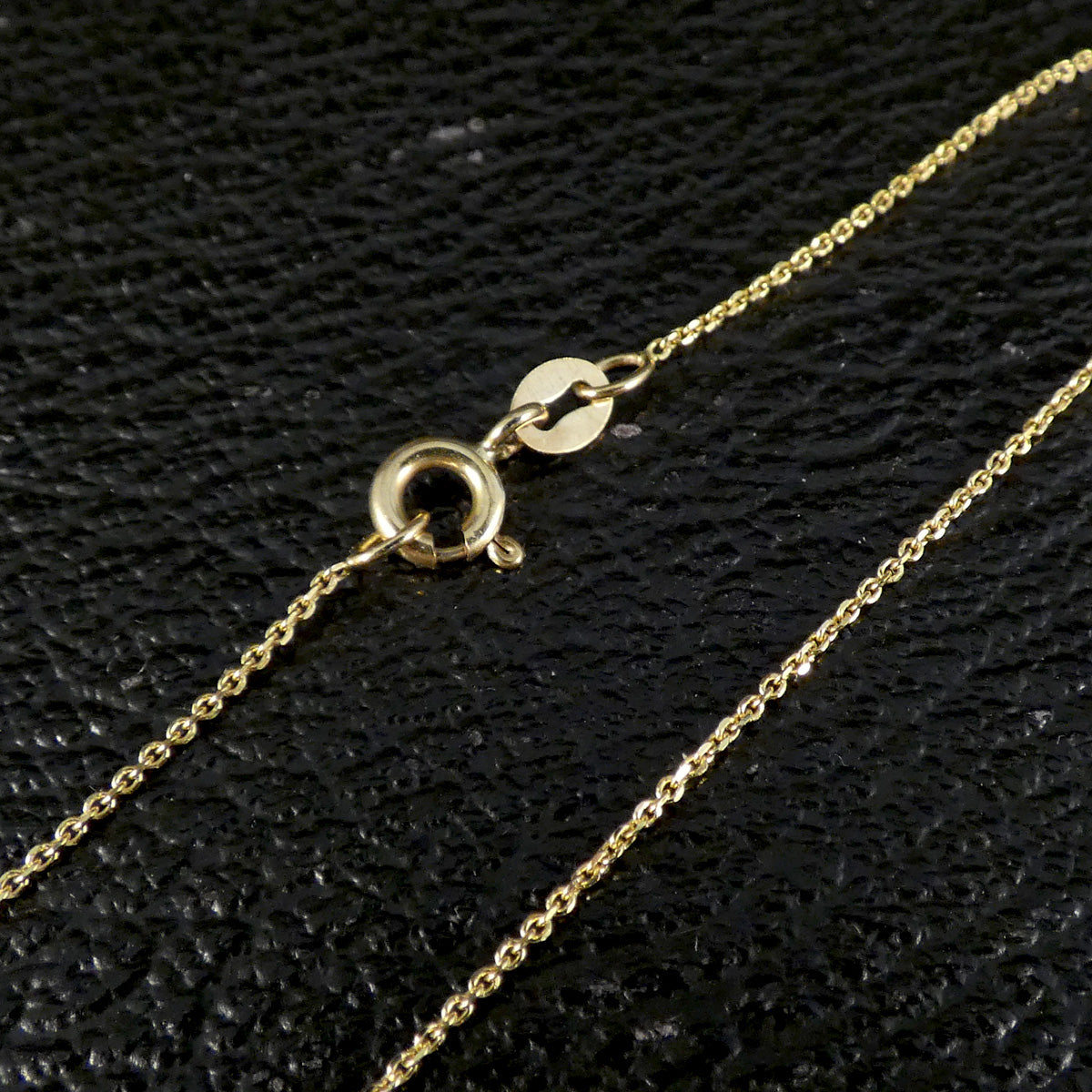 A close-up of a delicate gold chain necklace showing its lobster clasp and round tag, laid on a textured black surface.