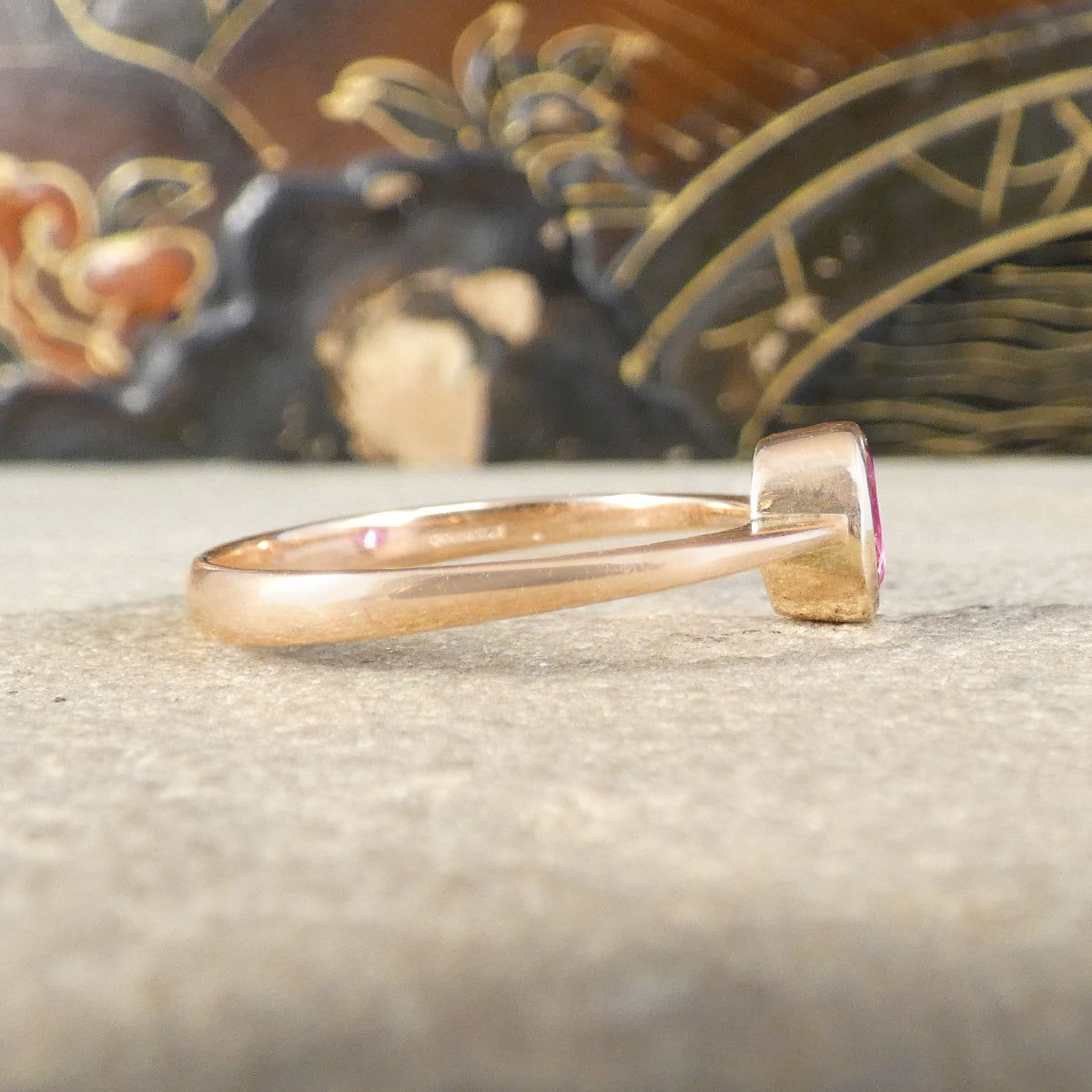 A close-up of a rose gold ring with a small pink gemstone, resting on a textured gray surface with a blurred decorative background.