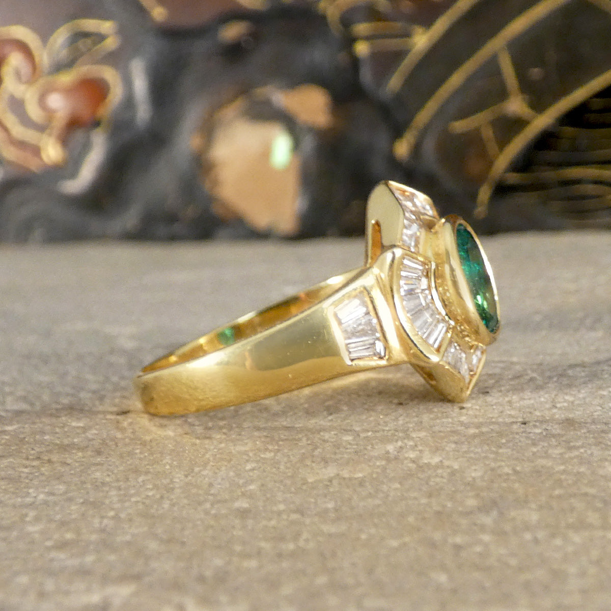 Side view of an emerald and tapered diamond cluster ring in 18ct yellow gold, showcasing a vibrant oval emerald centre stone with a halo of shimmering baguette diamonds set in a bold, elegant design.